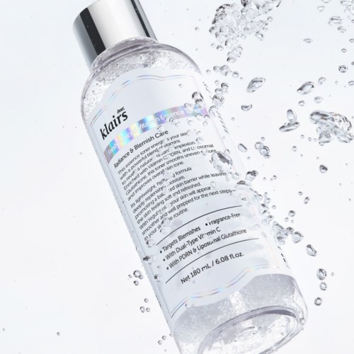 Eine klare Flasche mit Pumpe liegt auf einer weißen Oberfläche mit Wassertropfen. Das Etikett zeigt Hautpflegeinformationen.