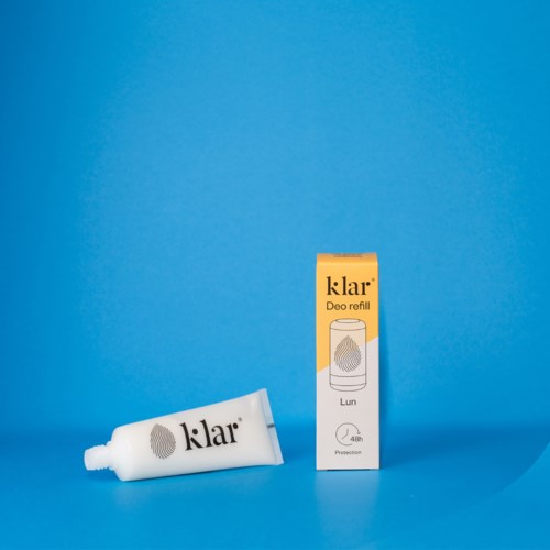 En hvit tube med Klar Deo refill og en gul eske med sprayikon på en blå bakgrunn.