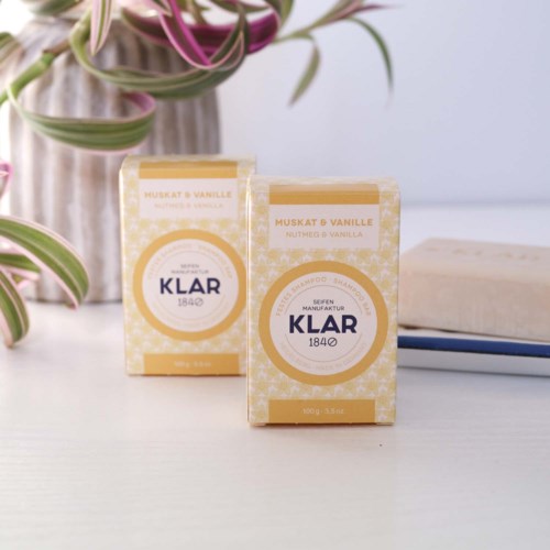 To små kasser mærket 'Klar 1840' med vanilje duft, placeret på en hvid overflade med planter og bøger tæt på.
