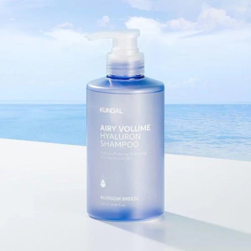 A blue shampoo bottle with a pump, labeled as "KUNDAL AIRY VOLUME HYALURON SHAMPOO." It is near water and sky.