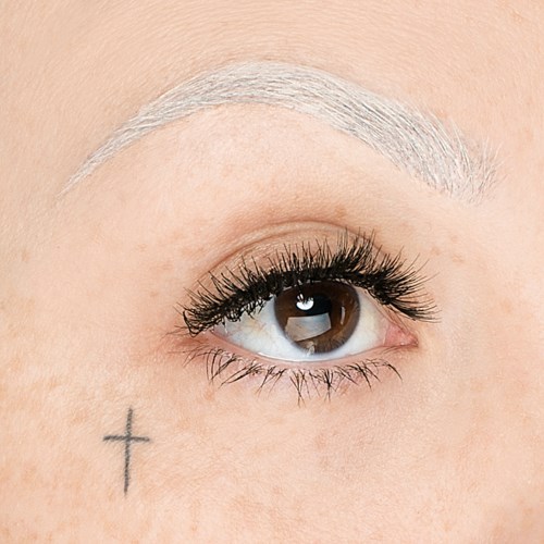 En nära bild av en persons öga med långa ögonfransar, vit ögonbryn och en liten kors tatuering på huden.