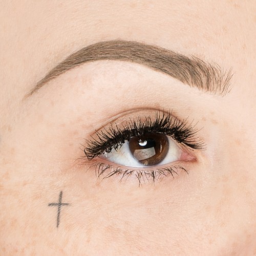 Närbild av ett öga med make-up, en liten tatuering av ett kors under ögat och väldefinierade ögonbryn.