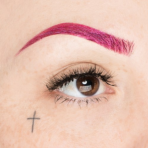 Närbild av en person med brunt öga, rosa ögonbrynsmakeup och en liten kors-tatuering under ögat.