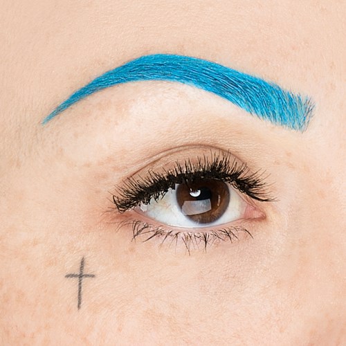 Närbild av ett öga med blå ögonbryn och en liten kors-tatuering i närheten.