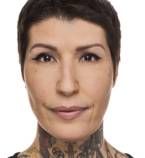 En kvinna med kort mörkt hår, tatueringar på nacken och smink, tittar direkt mot kameran.