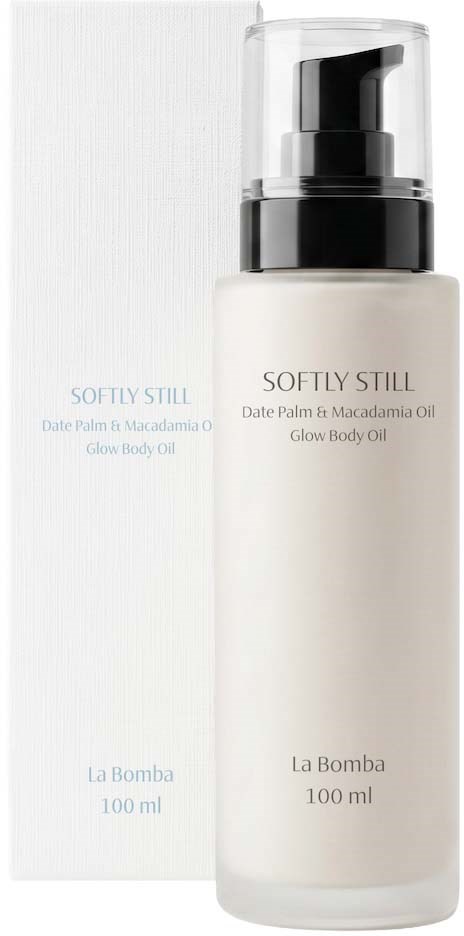 Biała butelka z czarnym dozownikiem i przezroczystym przykryciem, oznaczona Softly Still glow body oil od La Bomba.