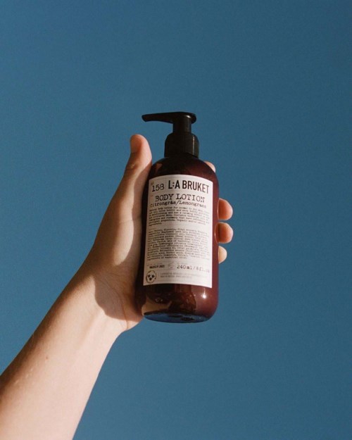 En hånd holder en brun flaske med body lotion og pumpe, mot en enkel blå bakgrunn.