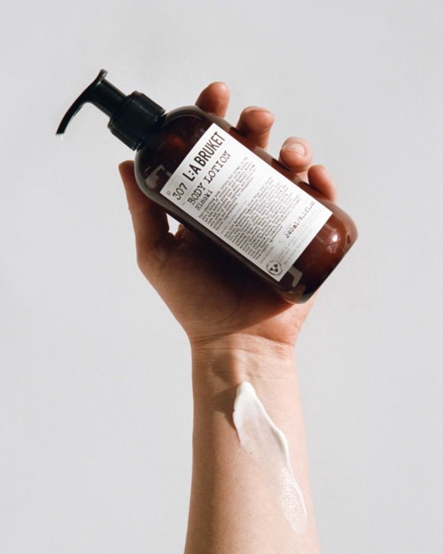 Eine Hand hält eine braune Flasche mit dem Etikett 'LA BRUKE BODY LOTION' vor einem schlichten Hintergrund.