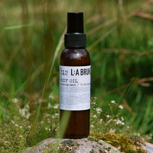 En liten brun sprayflaska märk med 'L:A BRUK' med kroppsolja, placerad utomhus på en sten med gräs i bakgrunden.