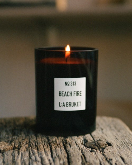 A black candle with a flame, labeled 'Beach Fire.' It sits on a rough wooden surface.