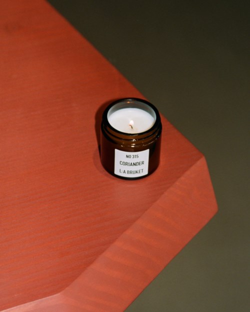 Een kleine bruine kaars in een pot met label "No. 35 Coriander La Bruket" op een houten oppervlak met schaduw.