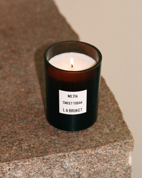 A black candle jar with a lit white candle on a stone surface. Label reads "NO 316 SWEET TOBAK L:A BRUKET."