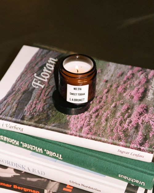 A small brown candle jar with a lit candle on top of stacked books, including one titled 'Torleif Rindal' and 'Ursula Nordström.'