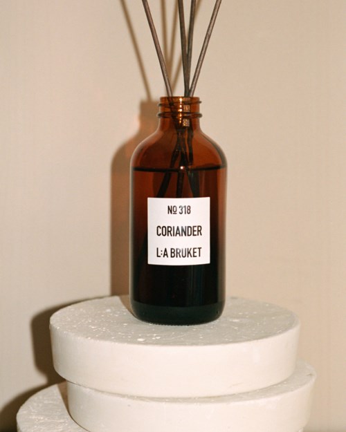 A brown bottle with reed sticks on a white round stand, labeled 'Coriander La Bruket', against a beige background.