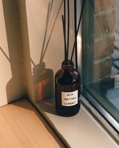 Brown bottle with black reed sticks on a windowsill, label says "Sweet Tobak."
