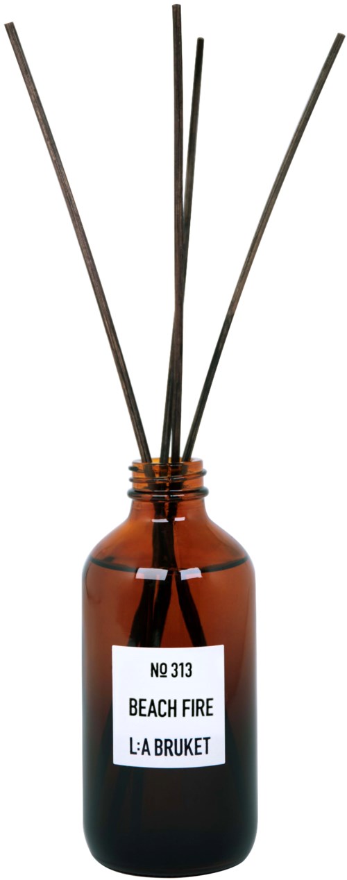 A brown glass bottle with a label and five thin sticks sticking out. The label reads "BEACH FIRE" and "L:A BRUKET."