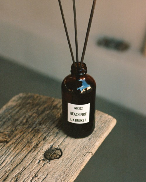 A small brown bottle with a label that says 'Beach Fire' and a person's name, with three sticks inside. It sits on a wooden surface.