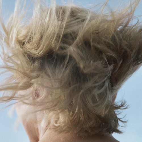 A person with messy, wind-blown blond hair, face partially visible, against a simple background.