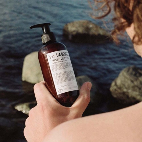 Een hand houdt een bruine fles bodylotion bij het strand, vlakbij stenen en water.