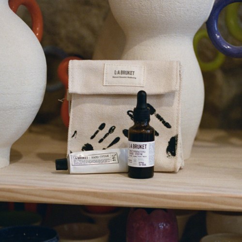 A small pouch and bottle of hand sanitizer from La Bruket sit on a wooden surface. The pouch has black script and images.