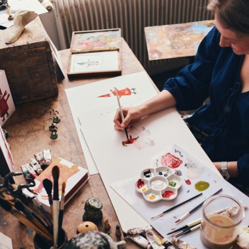 En kunstner maler på akvarelpapir med en palet af farver og pensler, omgivet af kunstmaterialer og skitser.