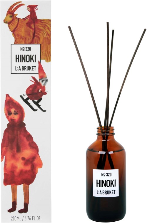 En brun glasflaske med sorte diffuserstænger og etiket, der siger Hinoki L:A Bruket, 320. Flasken står ved siden af dens emballage med farverige dyr og en person illustreret.