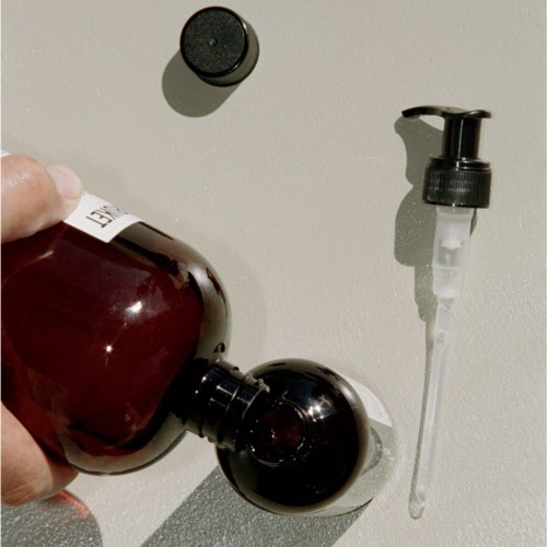 A person holds a brown bottle near a wall with a soap dispenser and a small black cap.