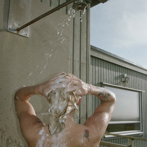 Person washing hair outdoors with water from a shower, next to a gray building.