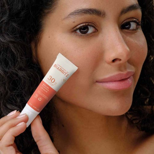 A woman with curly hair holds a sunscreen tube near her face, smiling slightly. No other objects are visible.
