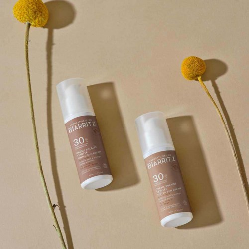 Zwei Flaschen Biarritz Sonnencreme mit SPF 30 sind neben gelben Blumen auf einem beigen Hintergrund platziert.