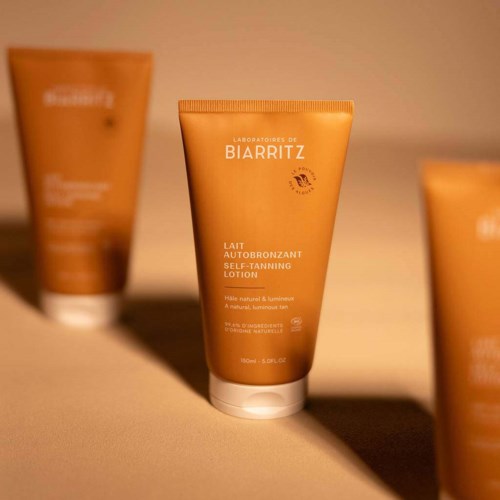 Orangener Tubensatz mit weißem Text, gekennzeichnet als Selbstbräunungslotion von Biarritz, mit Sonnenlichtthema. Zwei ähnliche Tuben im Hintergrund.