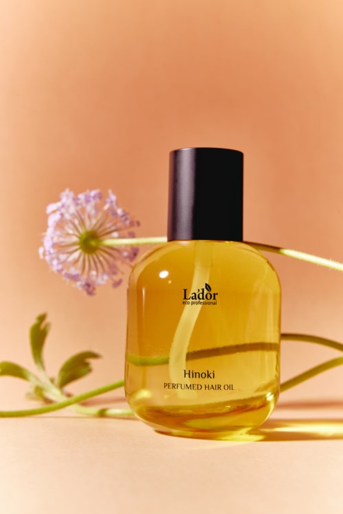Keltainen pullo hiusöljyä, jossa lukee 'Hinoki Perfumed Hair Oil', taustalla pinkki kukka ja vihreä kasvi.
