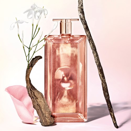 Eine rosafarbene Parfümflasche, umgeben von Blumenblättern, einem getrockneten Zweig und einem gewundenen Zweig, vor einem weichen Hintergrund.