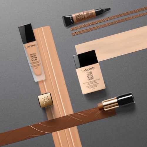 Makeup-produkter, inklusive foundation och concealer, är placerade med makeupborstar på en grå yta.