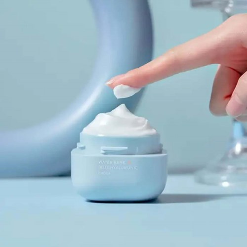 A person presses a finger into a jar of white cream on a light blue surface.