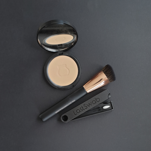 En makeupbørste, et kompakt pudder og et sort værktøj med et hul, alle på en mørk overflade.
