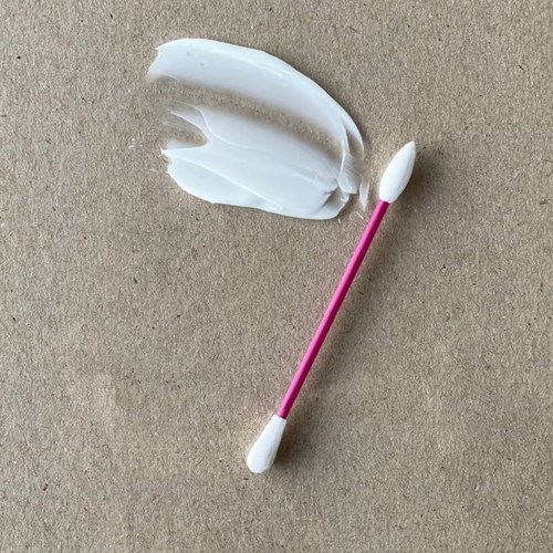 En lyserød og hvid vatpind ligger på en enkel beige overflade, med cremet makeup eller lotion smurt i nærheden.