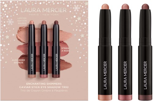 Tre svarta Laura Mercier ögonskuggsstift och deras förpackning med en ljus rosa bakgrund och snöflingedesign.