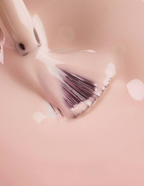 A rose gold makeup brush with white foam on it lies on a pink surface.