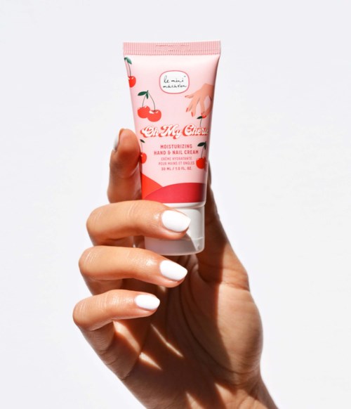 A hand holding a pink tube of moisturizing hand and nail cream with cherry illustrations.