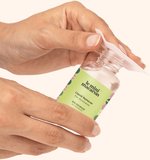 Hænder holder en lille gennemsigtig flaske med et grønt mærke, åbnet, mærket "le mini macaroon" og "liquid remover."