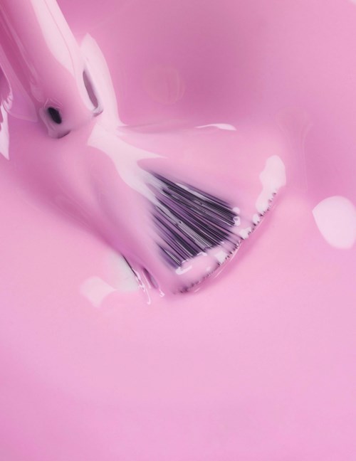 A makeup brush spreads pink cream or paint on a smooth surface. The brush has dark bristles and a pink handle.