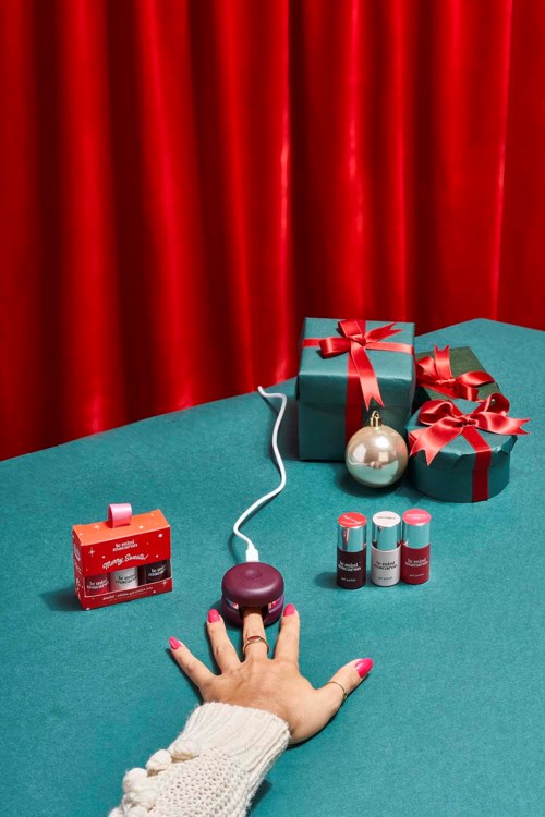 A hand with pink nails touches a device connected with a cord, surrounded by gift boxes and holiday decorations.