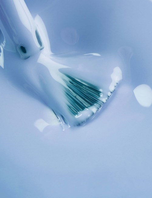 A paintbrush is submerged in blue paint, creating a smooth, flowing surface with some streaks visible.