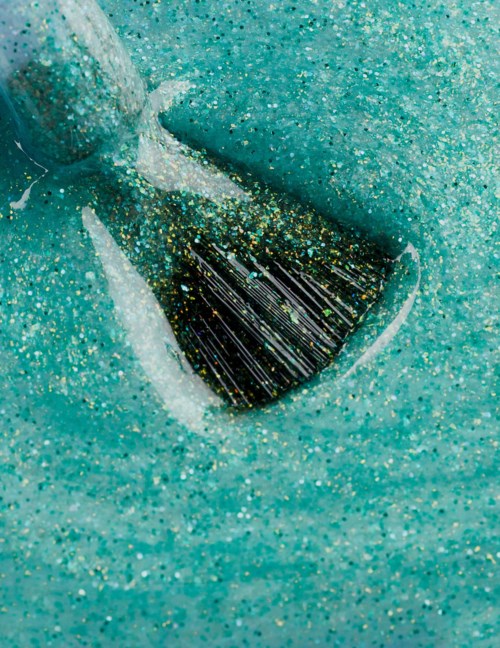 A small black object with glittering gold and green flecks is partially hidden in light blue sand.
