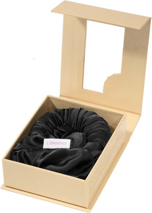 En cremefarvet kasse med et sort satin scrunchie indeni, og et gennemsigtigt vindue på låget.