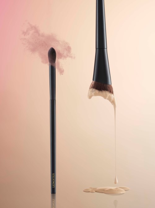 Två makeupborstar visas, en med brunt puder och den andra med flytande foundation som droppar.