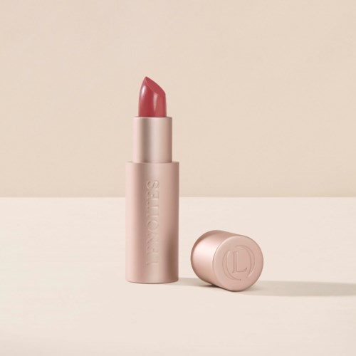 En rosa läppstift i en ljusrosa tub, med korken bredvid, på en beige bakgrund.