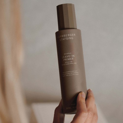 A hand holds a beige spray bottle labeled 'LERNBERGER STAFSING' with 'Leave-In Primer' written on it.