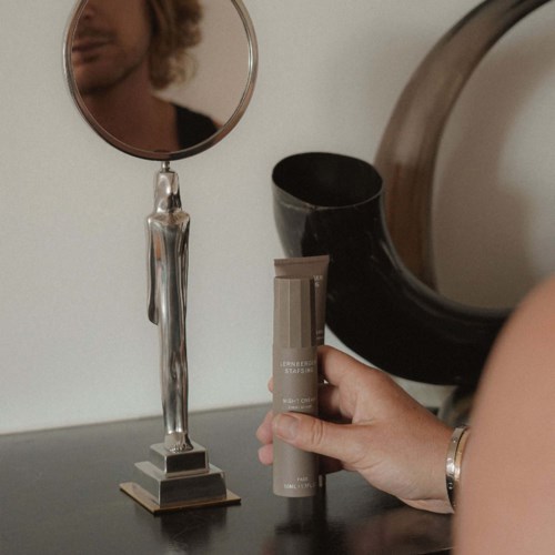 A person holds a skincare product near a mirror, with a black vase and decorative objects on the table.